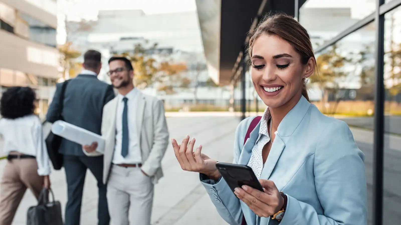 Collaboratrice consultant son smartphone devant des locaux d'entreprise, collègues en arrière-plan